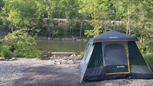 A camping tent by a river, surrounded by trees and a picnic table nearby on a gravel area.