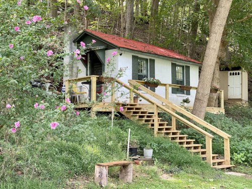 A small white cottage with a red roof, surrounded by greenery and flowers, featuring a wooden staircase and porch.