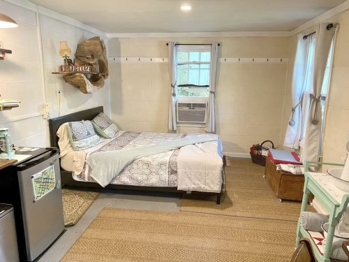 Cozy bedroom with a bed, air conditioner, small fridge, and natural decor, featuring light-colored walls and a woven rug.