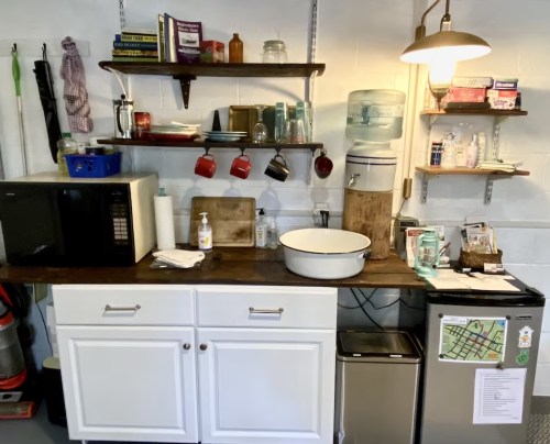 A cozy kitchen area with shelves, a microwave, water cooler, and various kitchen items on a wooden countertop.