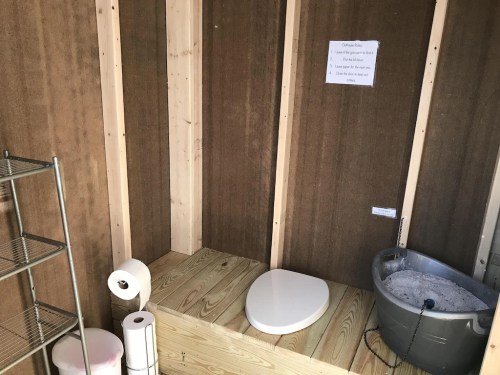A simple wooden bathroom setup with a compost toilet, toilet paper, and a storage shelf against a rustic wall.