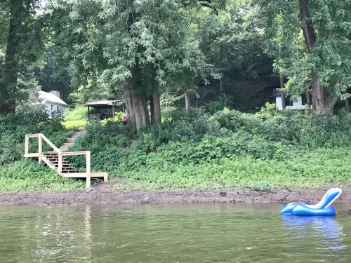 A serene river scene featuring wooden stairs leading to a house, with lush greenery and a blue inflatable float nearby.