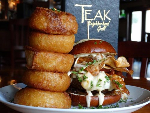 A stacked plate of crispy onion rings next to a gourmet burger topped with chips and creamy sauce, with a menu in the background.