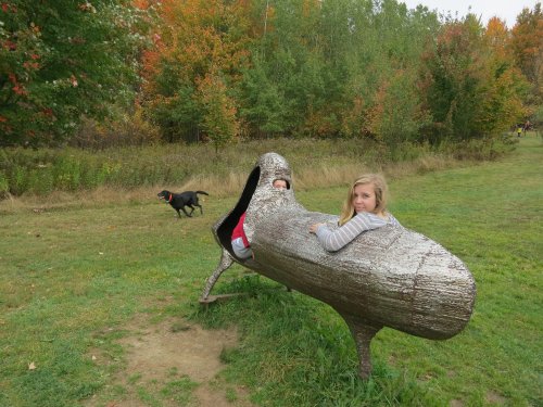 Two children sit in a large, whimsical sculpture resembling a shoe, with a dog running in the background and autumn trees nearby.
