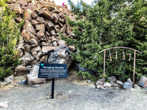 Sign for "Ringing Rocks" next to a rock formation and a metal arch with hanging bells in a natural setting.