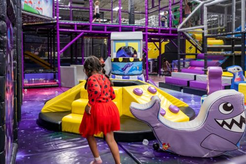 A child in a red dress plays in a colorful indoor playground with slides and climbing structures.