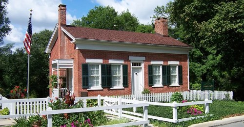 A historic brick house with white shutters, surrounded by a white picket fence and colorful flowers, under a blue sky.