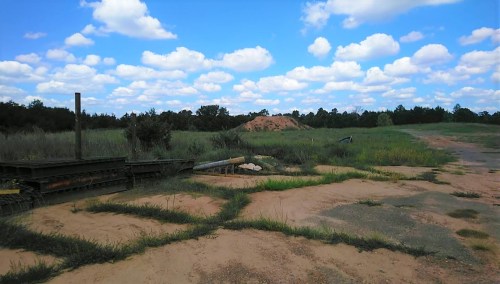 A grassy field with scattered clouds in a blue sky, featuring a dirt mound and some construction materials in the foreground.