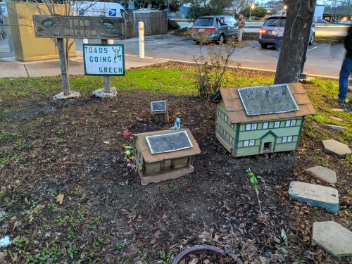 A whimsical garden scene featuring miniature houses and a sign reading "Toad Hollow: Toads Going Green."