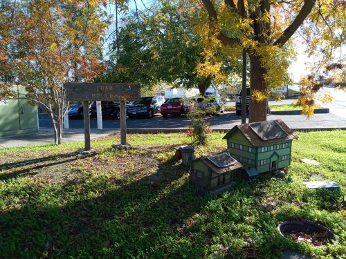 A small model house and sign in a park, surrounded by green grass and autumn trees with colorful leaves.