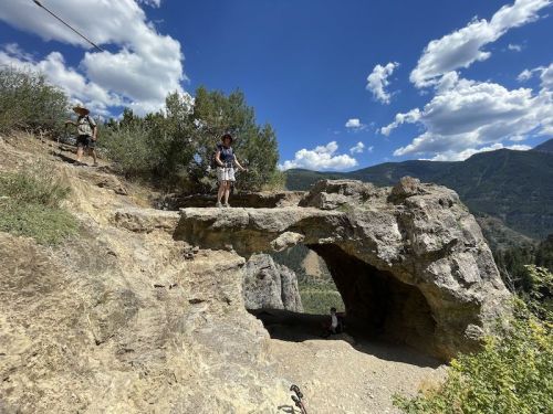 Two people explore a rocky landscape with a natural stone arch under a blue sky with fluffy clouds.