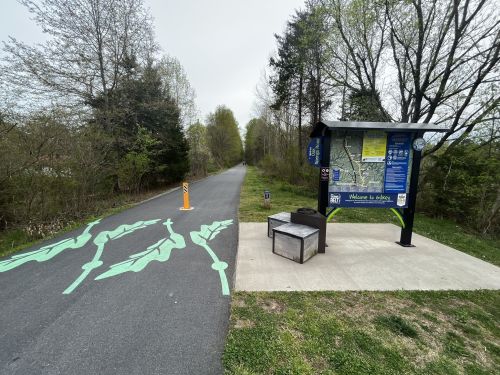 A trail with a map kiosk and green leaf designs on the path, surrounded by trees and greenery.