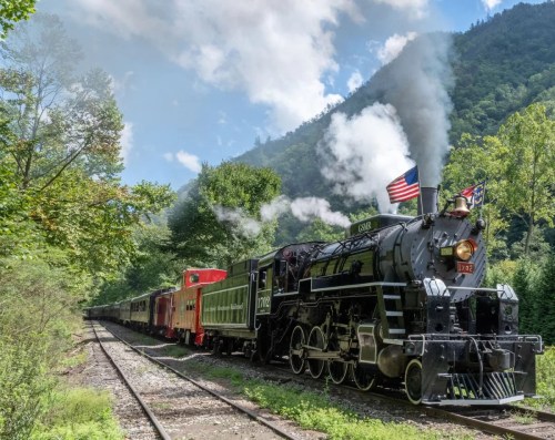 A vintage steam locomotive with smoke billowing, surrounded by lush greenery and mountains under a blue sky.