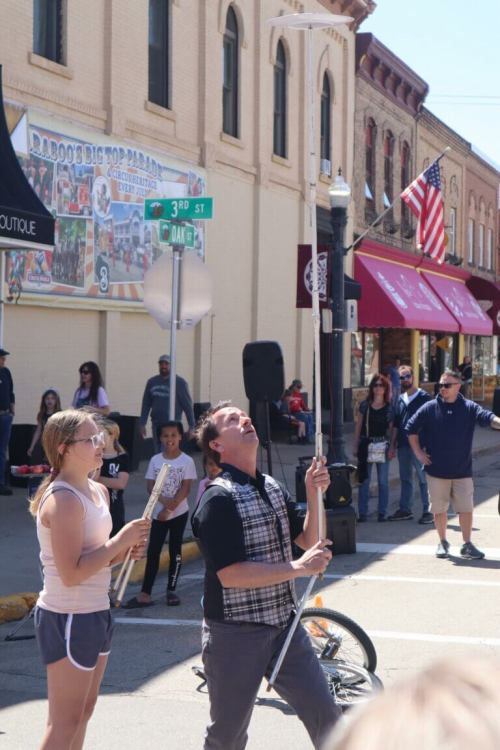 A street performer juggles while a woman assists, with a crowd watching in a lively downtown setting.