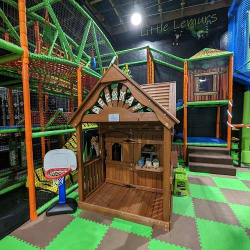 A colorful indoor play area featuring a wooden playhouse, climbing structures, and a basketball hoop.