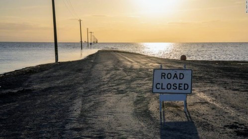 A "Road Closed" sign on a flooded road at sunset, with power lines visible in the background.