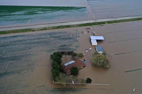 Aerial view of a flooded area with a house and outbuildings partially submerged in water, surrounded by fields.