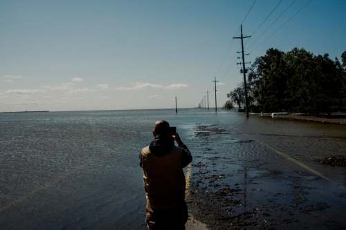 A person stands on a flooded road, taking photos of submerged power lines and trees under a clear blue sky.
