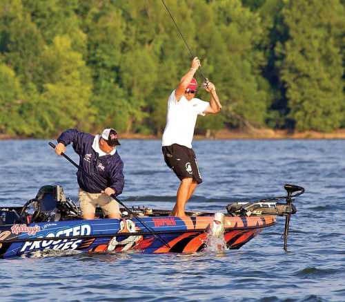 Two men fishing from a boat, one reeling in a fish while the other assists, surrounded by lush greenery and water.