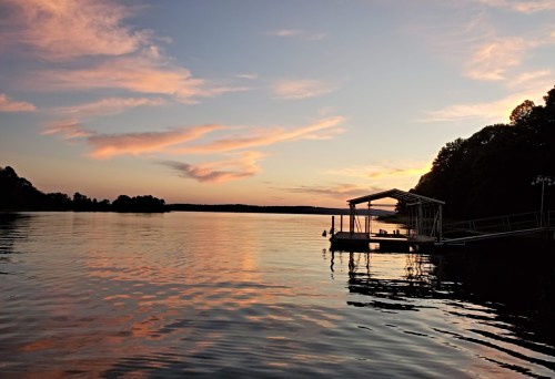 A serene lake at sunset, with colorful clouds reflecting on the water and a dock silhouetted against the horizon.