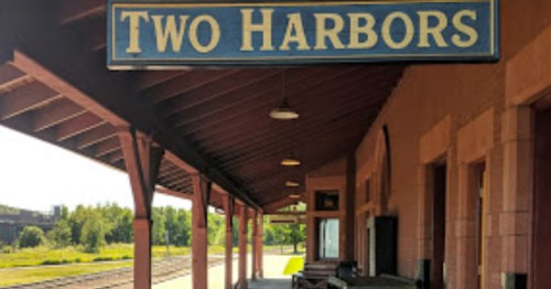 Sign reading "Two Harbors" at a train station with a covered platform and green landscape in the background.