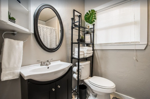 A modern bathroom featuring a round mirror, sink, toilet, and a black shelving unit with towels and plants.