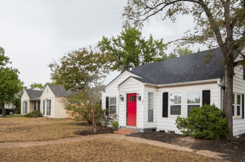 A white house with a red door, surrounded by greenery, alongside two other homes in a residential area.