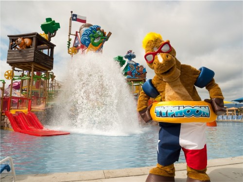 A mascot in sunglasses holds a floatie by a water park with slides and a splash area in the background.