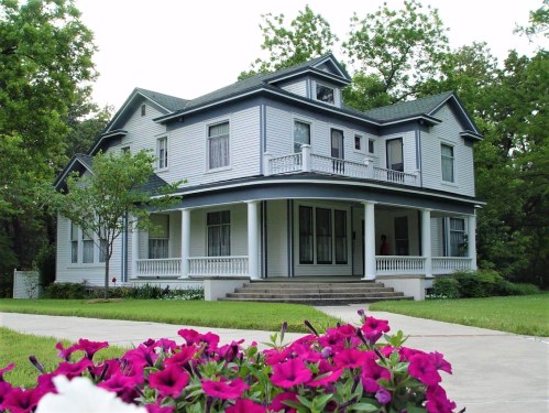 A large, two-story gray house with a wraparound porch, surrounded by green trees and colorful flowers in the foreground.