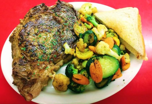 A plate featuring a grilled steak, mixed vegetables, and a slice of bread on a red table.