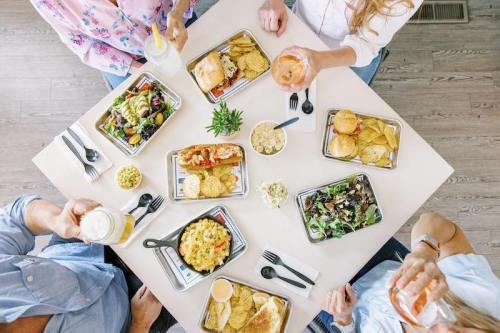 A group of people enjoying a variety of meals on trays at a table, with drinks and salads visible.
