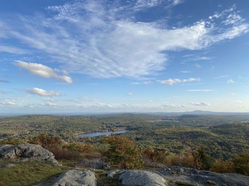 A scenic view of rolling hills and a lake under a blue sky with scattered clouds, showcasing autumn foliage.