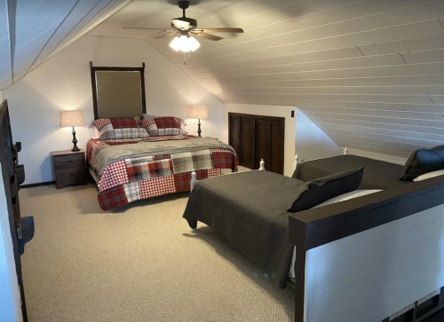Cozy attic bedroom with a queen bed, a twin bed, and warm lighting from lamps. White walls and sloped ceiling create a charming space.