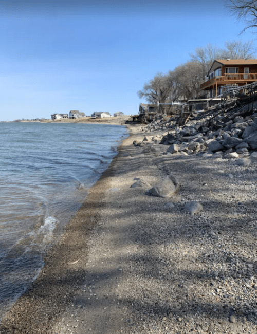 A serene lakeside view with a sandy shore, rocks, and houses in the background under a clear blue sky.