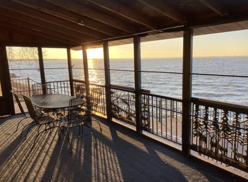 A sunlit porch with a table and chairs overlooking a calm lake at sunset, framed by screens and wooden beams.