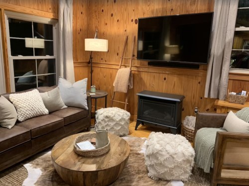 Cozy living room with wooden walls, a TV, a brown sofa, fluffy ottomans, and a round coffee table. Warm and inviting atmosphere.