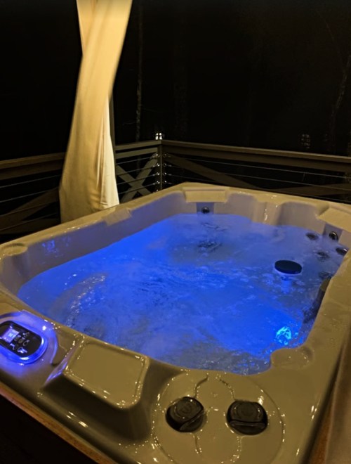 A lit hot tub with bubbling water, set on a deck at night, surrounded by a dark, wooded area.