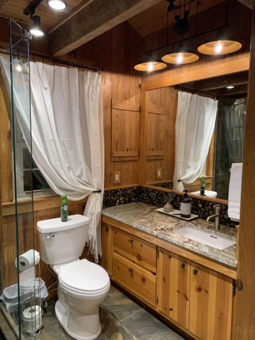 Cozy bathroom with wooden walls, a glass shower, and a granite countertop, featuring a toilet and mirror with warm lighting.