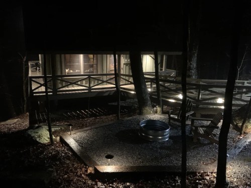 A dimly lit cabin at night, surrounded by trees, with a fire pit and outdoor seating area visible in the foreground.