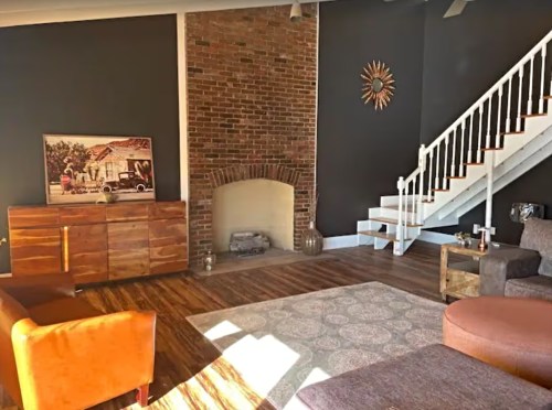 Cozy living room with a brick fireplace, wooden furniture, and a staircase, featuring warm lighting and a patterned rug.