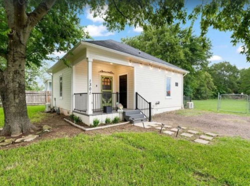 A charming white house with a black railing, surrounded by greenery and a gravel path, under a clear blue sky.