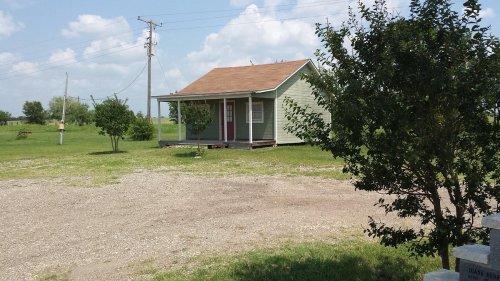 A small green house with a brown roof, surrounded by grass and trees, under a blue sky with scattered clouds.