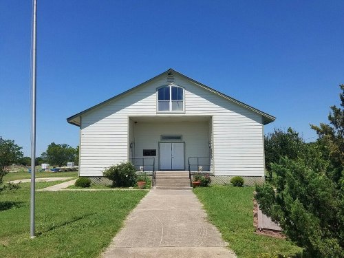 A white church building with a peaked roof, steps leading to the entrance, and greenery surrounding it under a clear blue sky.