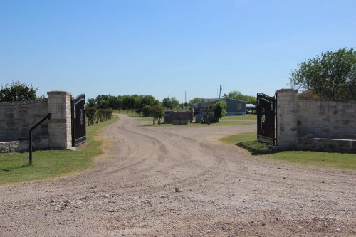A gravel driveway leads through open gates into a rural area with green trees and a blue building in the distance.
