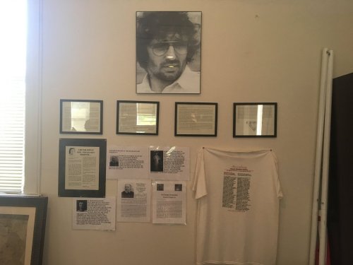 A wall display featuring a portrait, framed texts, and a shirt with names, all arranged in a gallery-like setting.