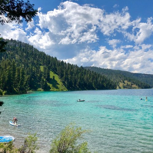 A serene lake surrounded by lush green hills, with people paddleboarding and kayaking under a partly cloudy sky.