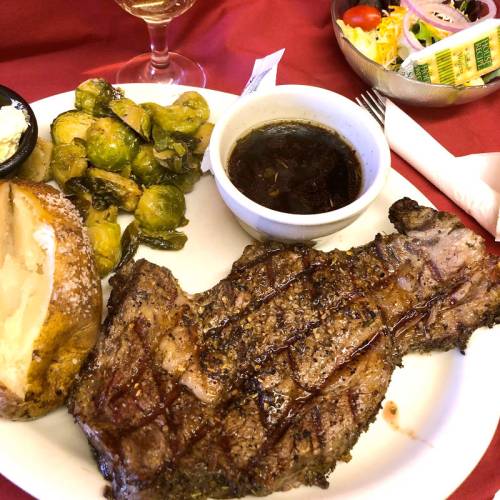 A plate featuring a grilled steak, Brussels sprouts, a baked potato, and dipping sauce, with a salad on the side.