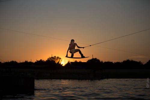 A person performs a wakeboarding jump against a sunset backdrop, silhouetted against the water and trees.