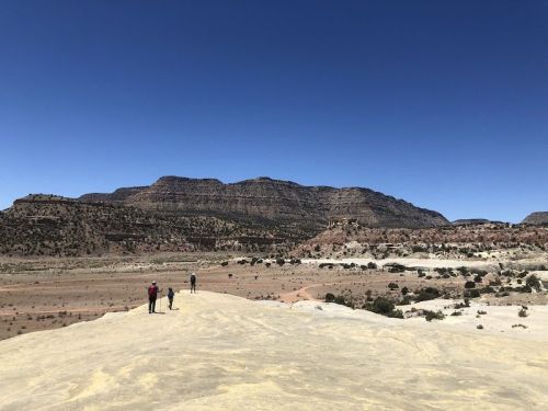 Two hikers walk along a rocky landscape under a clear blue sky, with mountains in the background.