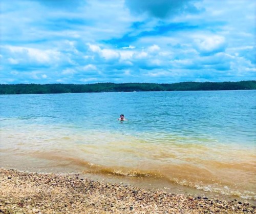 A person wades in a calm lake with a sandy shore under a cloudy blue sky. Green hills are visible in the background.